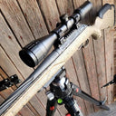 Thermal rifle scope mounted on camo rifle, set on tripod against wooden backdrop for predator hunting